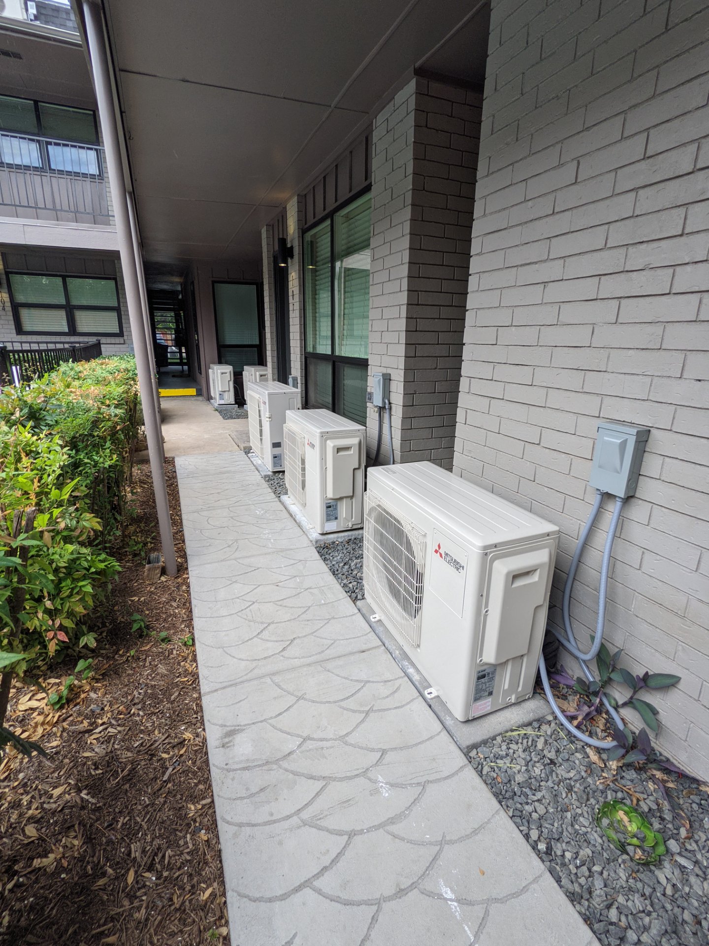 Multiple Mitsubishi Electric mini-split outdoor units installed on gravel beside a building walkway.