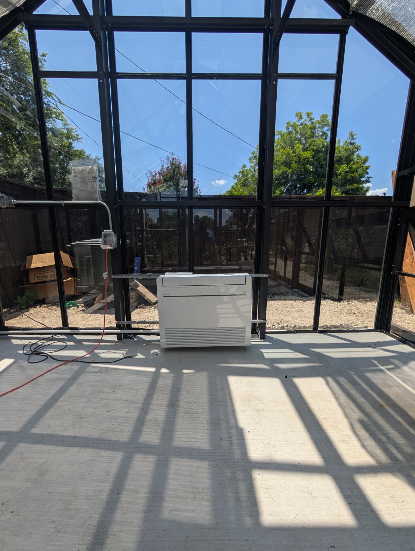 Mini-split indoor air handler installation in a sunroom with large glass panes on a concrete floor.