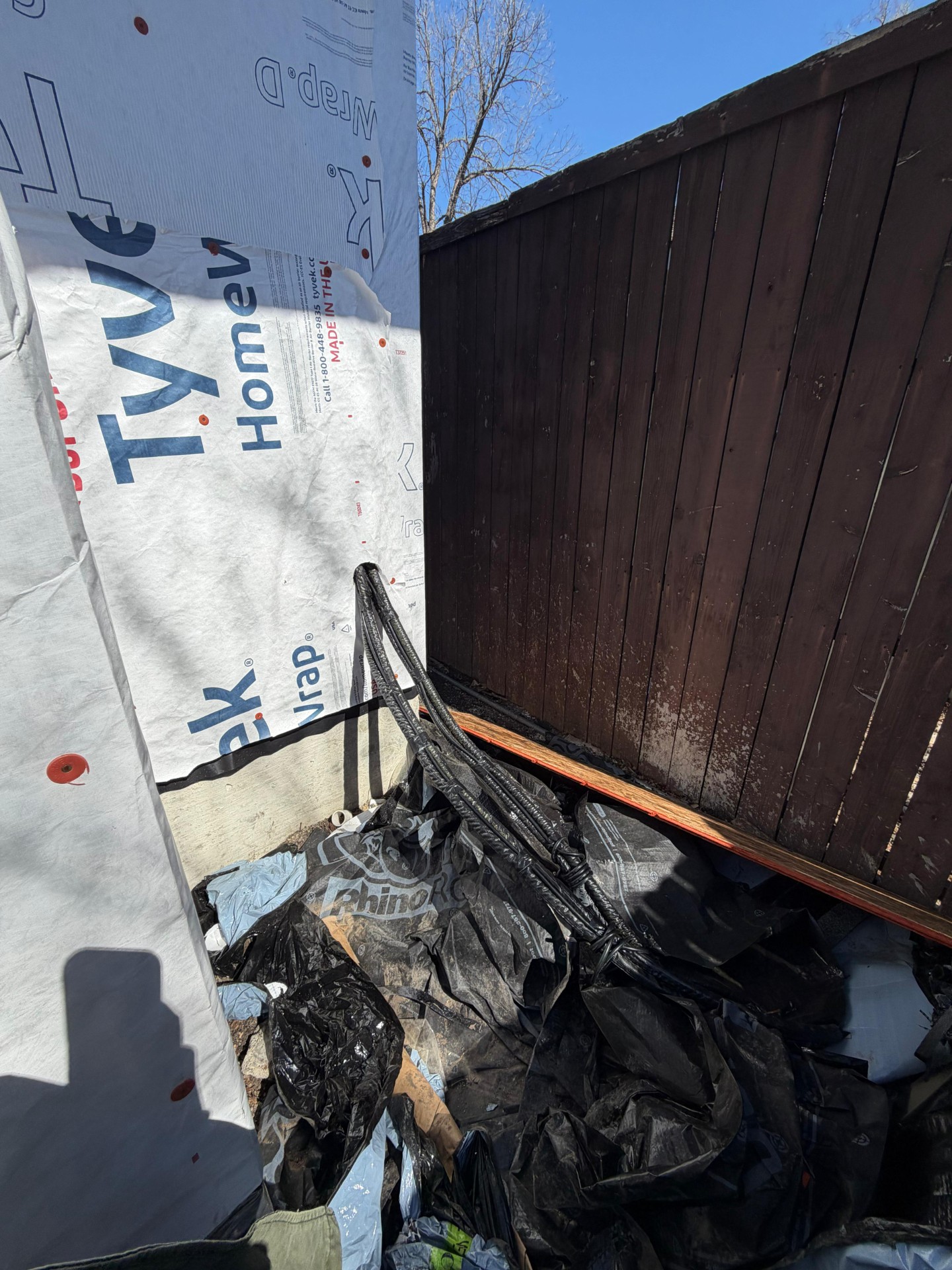 Construction wrap and discarded materials next to a wooden fence, indicating an ongoing HVAC or building project.