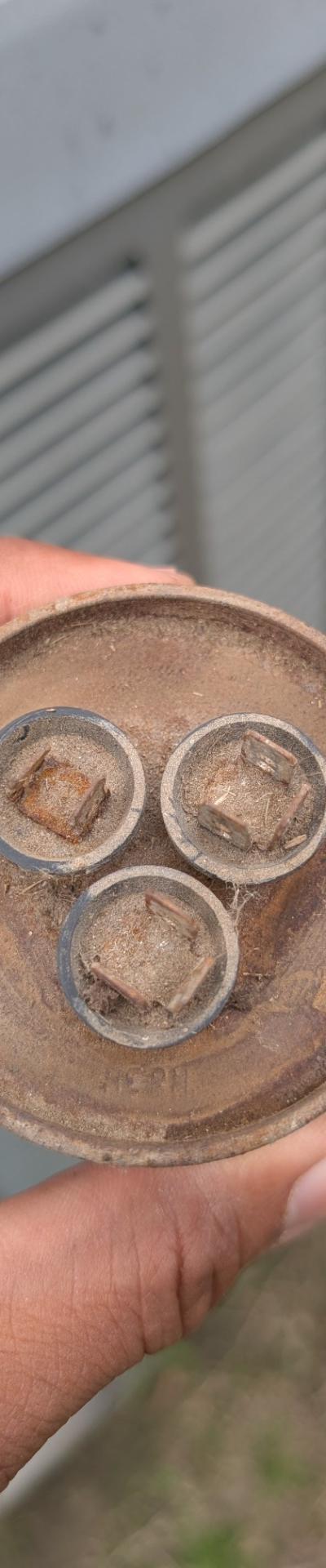 Technician holding a dirty, corroded HVAC capacitor in front of an outdoor unit, signaling a repair or replacement.